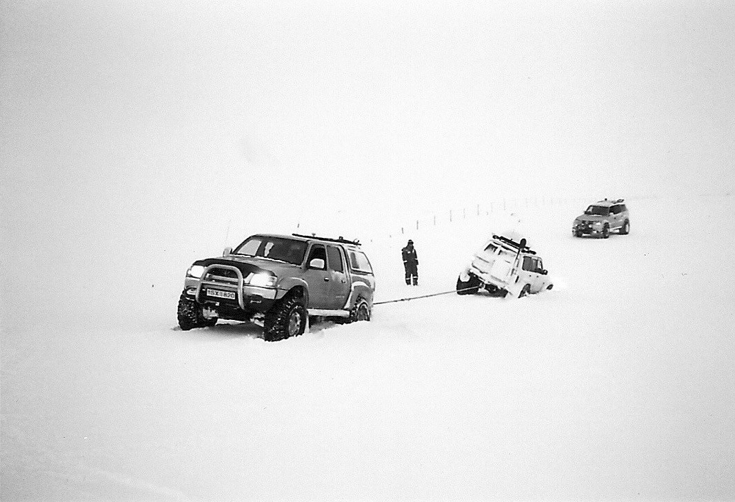 Paysage de neige islandais. Une voiture est visiblelment bloquée, et une autre la tire.