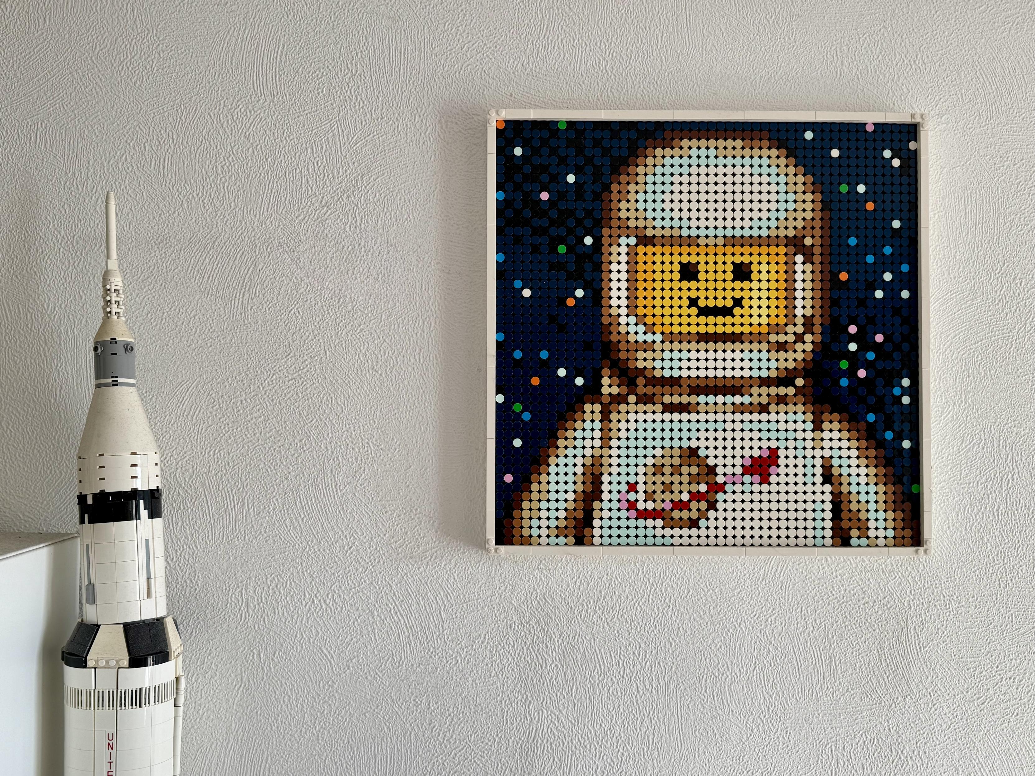 Un cadre lego représentant une Minifig astronaute devant l’espace étoilé. Le cadre est sur un mur et on aperçoit aussi le haut d’un statue Saturn V.