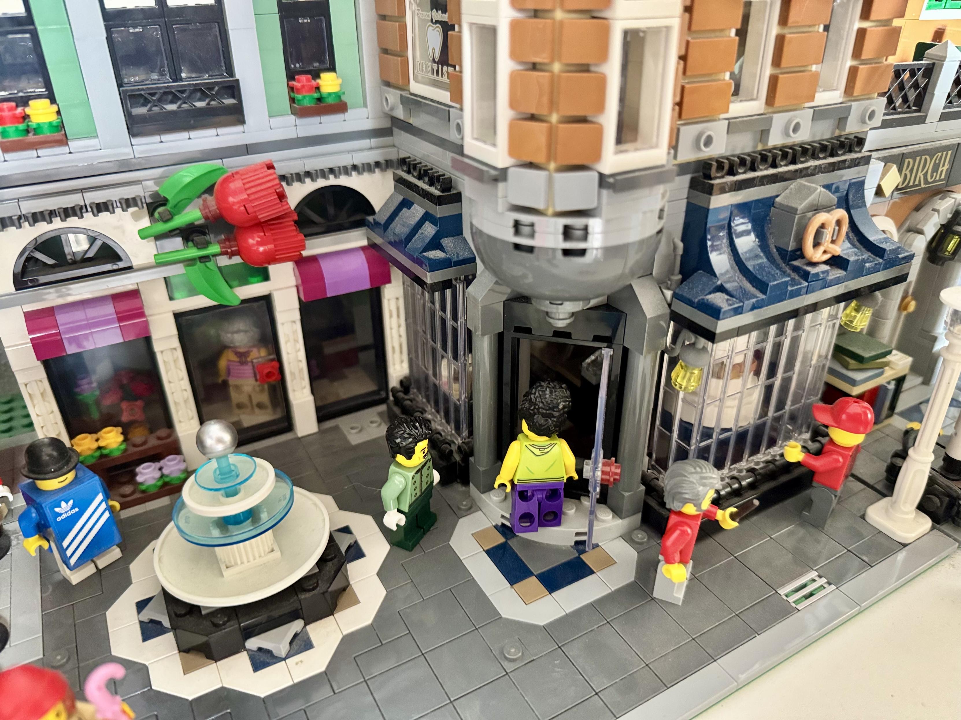 Sur cette place lego, autour d’une fontaine, se trouve un magasin de fleurs et une boulangerie.